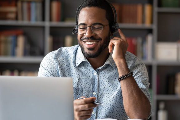 Smiling African American man in glasses and headset watch webinar on laptop making notes, happy biracial male student worker in headphones handwriting studying or working using computer